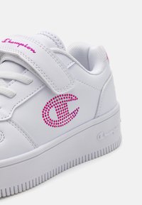 Baskets blanches avec une tige en synthétique lisse, une sangle velcro, une semelle plateforme surélevée et un logo en rhinestones roses sur le côté.