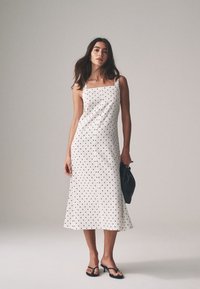 White polka dot dress with a square neckline and thin straps. Flared hem and smooth fabric, paired with black accessories and sandals.