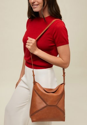 Punases lühikeste varrukatega topis ja valgetes pükstes naine kannab pruuni nahast õlakotti geomeetrilise õmblusdetailidega.