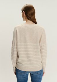Beige trui van zacht, gestructureerd materiaal, met horizontale witte strepen. Overmaat ontwerp met verlaagde schouders en geribde zoom.