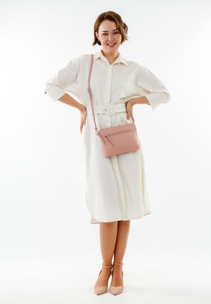 Robe chemise blanche avec une taille cintrée, associée à un petit sac bandoulière en cuir rose. Le mannequin porte des talons nus, mettant en valeur des textures lisses.