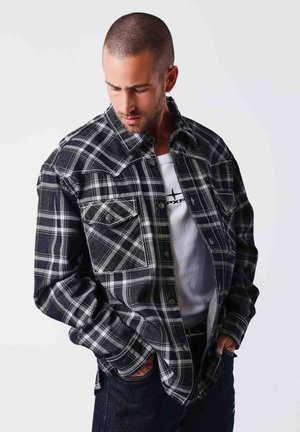 Homme aux cheveux courts portant une chemise à carreaux noir et blanc par-dessus un t-shirt blanc à motif, regardant vers le bas, mains dans les poches, sur fond uni.