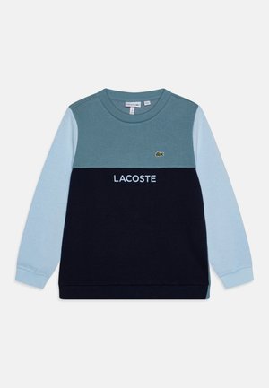 Bluza z długim rękawem i okrągłym dekoltem, z jasnoniebieskimi rękawami, morskim górnym fragmentem, czarną dolną częścią, tekstem "LACOSTE" i logo krokodyla na piersi.