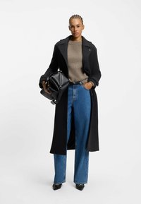 Zwarte wollen jas met een lange ontwerp, gedragen over een beige gebreide trui en blauwe wijde jeans. Houdt een zwarte tas vast. Punttip zwarte schoenen.