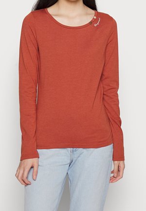 Vrouw draagt een langarmshirt in roestoranje met knopen en een klein wit schriftdetail bij de halslijn, gecombineerd met lichtblauwe jeans.