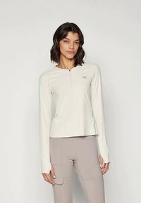 Light beige long-sleeve top with a zippered neck, smooth texture, flexible fit, and a small logo on the chest, paired with light pants.