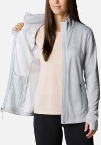 Hellgraue Fleece-Jacke mit durchgehendem Reißverschluss, strukturiertem Innenfutter, seitlicher Reißverschlusstasche und Stehkragen, getragen über einem rosa Oberteil.