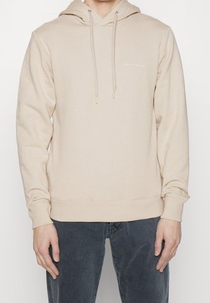 Personne portant un sweat à capuche beige clair avec cordons et un petit texte "Tiger of Sweden" sur la poitrine, associé à un jean délavé foncé.