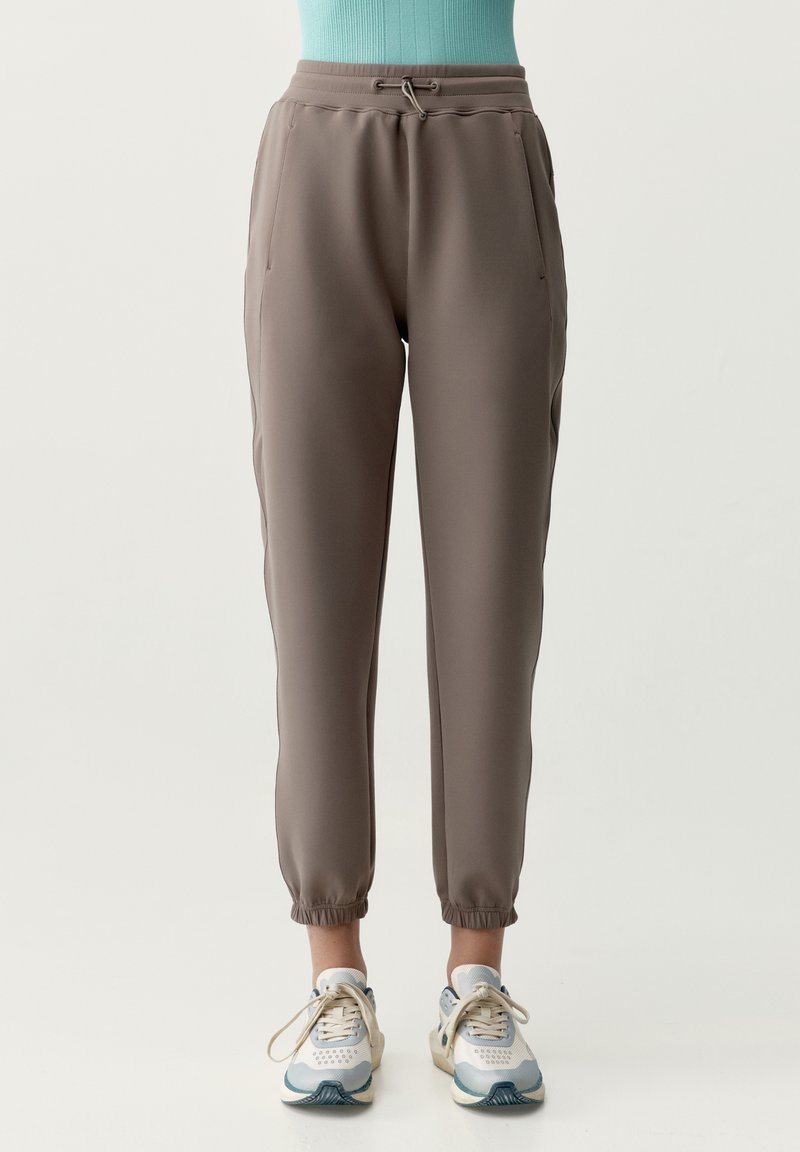 Taupe taps toelopende joggerbroek met elastische boorden, zijzakken en een taille met trekkoord, gedragen met witte en blauwe sneakers.