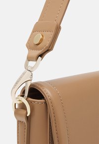 Detalj av en tan-färgad läderväska med en guldfärgad klämma och hårdvara, med eleganta linjer och synlig söm längs kanterna.