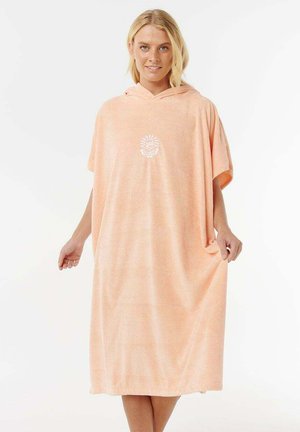 Donna bionda che indossa un vestito accappatoio con cappuccio largo color pesca con emblema bianco di sole e onda, in piedi davanti a uno sfondo bianco uniforme.