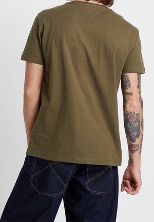 Homme portant un t-shirt vert olive et un jean bleu foncé, montrant des tatouages détaillés sur son bras droit, se tenant devant un fond uni.