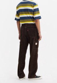 Pantalones de pana marrones con un corte relajado, que cuentan con bolsillos traseros y un parche de marca. Combinados con una camisa a rayas en azul marino, verde y blanco.