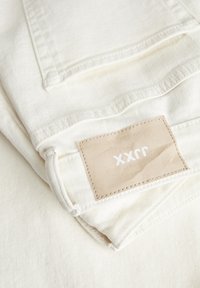 Tecido de ganga branca dobrado cuidadosamente, com uma etiqueta de couro bege com o texto "xx", costuras visíveis e uma textura levemente canelada.