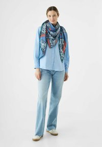 Chemise bleu à manches longues, jeans bleu clair, et une écharpe à motif présentant des tons de bleu, rouge et noir, avec une bordure à franges. Chaussures décontractées.