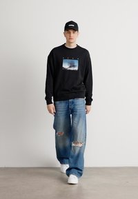 Felpa nera con un design grafico, abbinata a jeans strappati azzurri e sneakers bianche. Cappello con il logo "Hugo". Sfondo piatto e texturizzato.