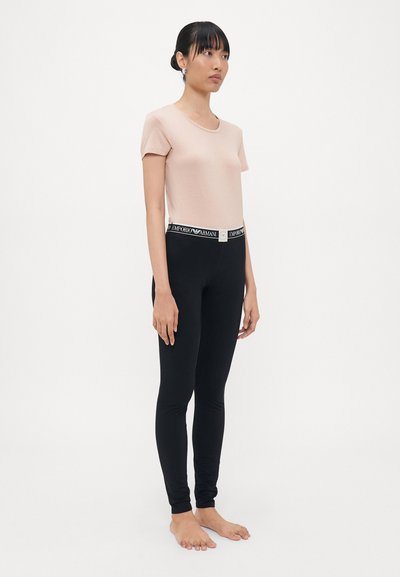 Kvinna som står barfota iklädd en ljusrosa t-skjorta med kort ärm och svarta leggings med en märkesbeteckning från Emporio Armani i midjan, mot en vit bakgrund.