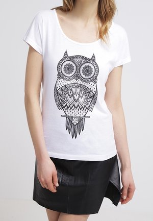 Femme portant un t-shirt blanc avec un motif détaillé de chouette noire, associé à une jupe en cuir noire asymétrique.