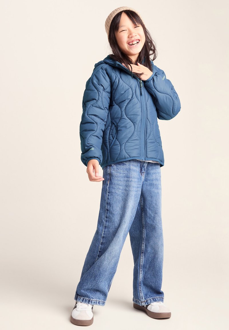 Smiling child wearing a beige beanie, blue quilted jacket, loose blue jeans, and white sneakers standing against a plain beige background.