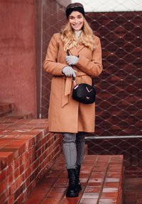 Brauner mantel mit Gürtel, graue Handschuhe, graue Hose und schwarze Kampfstiefel. Schwarze Umhängetasche mit gestepptem Muster und pinken Akzenten. Texturierter Hintergrund.