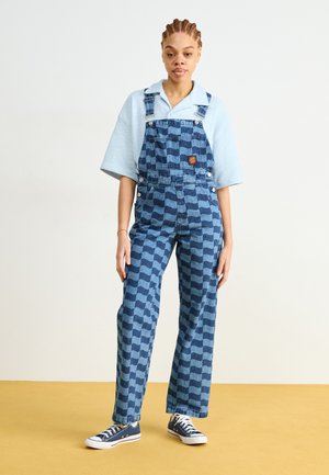 Mameluco de mezclilla con un patrón de cuadros azules, botones de metal y un parche. Combinado con una camiseta de manga corta azul clara y zapatillas navy.