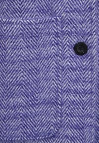 Látka s herringbone vzorem v purpurové barvě s měkkou texturou, s černým knoflíkem na pravé straně a náprsní kapsou.