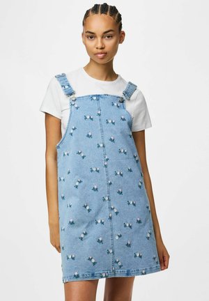 Jeune femme aux cheveux tressés porte un t-shirt blanc sous une robe en jean bleu clair avec de petites fleurs blanches et roses brodées.
