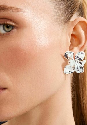 Orecchino a forma di fiore tono argento con superficie lucida e contorni organici, applicato a un orecchio testurizzato, che mette in risalto i dettagli nel metallo.