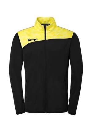 Veste noire zippée avec empiècement et col à motif géométrique jaune, portant le nom de la marque "Kempa" sur le côté gauche de la poitrine.
