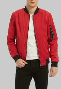 Veste bomber rouge avec une fermeture éclair à l'avant, des poignets et un ourlet en ribbed noir, et une poche latérale avec un détail en étiquette. Portée sur une chemise blanche et un pantalon noir.