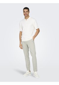 Camisa polo blanca de manga corta con cierre, combinada con pantalones slim-fit de color gris claro y zapatillas blancas, todos con texturas suaves y una silueta moderna.