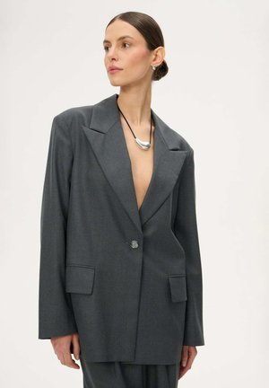 Kobieta z zaczesanymi do tyłu włosami nosi oversize'ową ciemnoszarą marynarkę i srebrny naszyjnik z wisiorkiem na jednolitym jasnym tle.