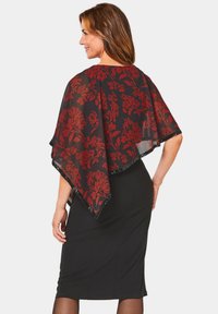 Poncho noir transparent avec un motif floral rouge et des accents en noir, porté par-dessus une robe noire ajustée jusqu'aux genoux. Le textile semble léger.