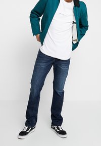 Giacca teal da uomo con colletto nero, t-shirt bianca con un design a linee, jeans blu scuro e sneakers nere con lacci bianchi.