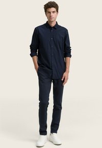 Uomo in piedi con le mani in tasca, indossa una camicia a bottoni blu navy, pantaloni blu navy abbinati e sneakers bianche, su uno sfondo beige uniforme.