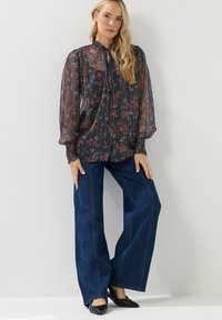 Durchsichtige schwarze Bluse mit Blumenmuster, hohem Kragen und langen Puffärmeln. Kombiniert mit weit geschnittenen blauen Jeans und schwarzen spitzen Ballerinas.