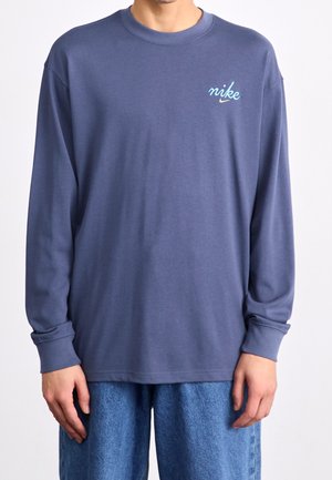 Niebieska koszulka z długim rękawem wykonana z bawełny, z okrągłym dekoltem i małym logo Nike w jasnoniebieskim kolorze na lewej piersi. Gładka faktura.