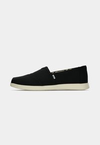 Chaussure noire sans lacets avec une semelle blanche, un petit talon, une partie supérieure élastique et une petite étiquette en tissu sur le côté.