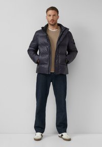 Tamna puffer jakna s visokim ovratnikom, s patentnim zatvaračem i bočnim džepovima, nosi se preko bež pletene veste, u kombinaciji s tamnim trapericama i tenisicama.