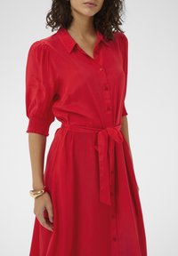 Robe rouge boutonnée avec col, manches courtes bouffantes et taille nouée. Texture lisse, longueur genou, avec une coupe décontractée et un léger éclat.