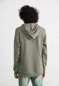 Felpa verde oliva con una texture morbida e un cappuccio con coulisse. Presenta maniche lunghe e una vestibilità rilassata, abbinata a jeans azzurri.