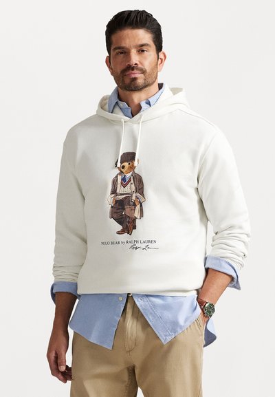 Mand iført en hvid hoodie med et Polo Bear grafik over en blå skjorte og beige bukser, stående med den ene hånd i lommen.