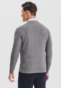 Mężczyzna odwr�ócony plecami, ubrany w szary sweter na wzorzystą białą koszulę i ciemne spodnie, na jednolitym białym tle.