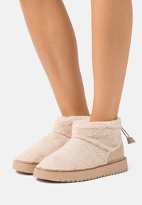 Beige ankle boots featuring a fluffy exterior, smooth leather accents, and a rubber sole with textured patterns. Drawstring detail at the back.