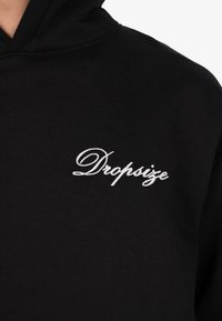 Czarna bluza z kapturem z białym, kursywą napisem "Dropsize" wyszytym na lewej części klatki piersiowej, ukazująca część ramienia i kaptura.