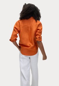 Oranje linnen overhemd met opgerolde mouwen, voorzien van een rugplooitje en een ronde zoom, gecombineerd met witte linnen broek. Schoon en minimalistisch ontwerp.