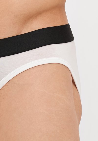 Weiße Baumwollunterhose mit einem schwarzen elastischen Bund, die eine glatte Textur und einen tiefen Schnitt an den Seiten aufweist.