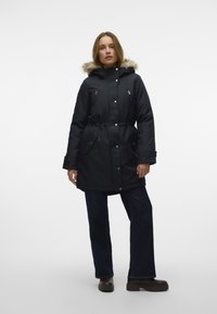 Melns parka mētelis ar mākslīgā kažoka kapuci, sašaurinātu jostu un ar rāvējslēdzējiem aizdarām kabatām. Ietver pogu aizdari un vienkāršu auduma struktūru.