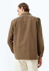 Brun kanvas skjortjacka med knäppt krage, långa ärmar och sidofickor. Har en subtil struktur och en skräddarsydd passform, bärs med ljusa byxor.