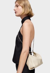 Top negro sin mangas con cuello alto, diseño sin espalda. Bolso beige claro con cadena, textura suave y brillo sutil.